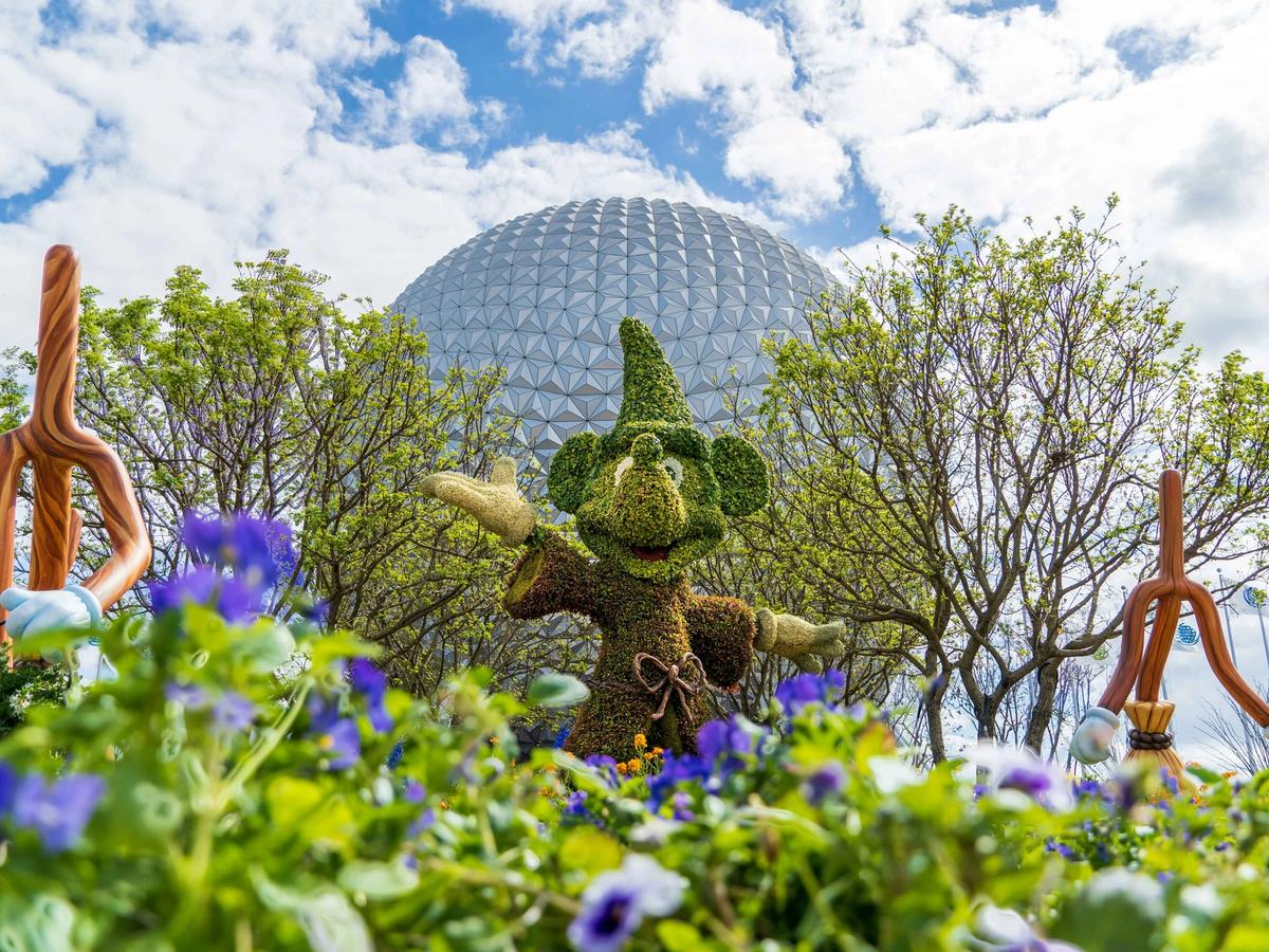A colorful Mickey Mouse topiary dressed as the Sorcerer stands in front of EPCOT’s famous geosphere, surrounded by blooming flowers and spring trees. It’s a picture-perfect day at EPCOT, full of fun, imagination, and Disney charm.