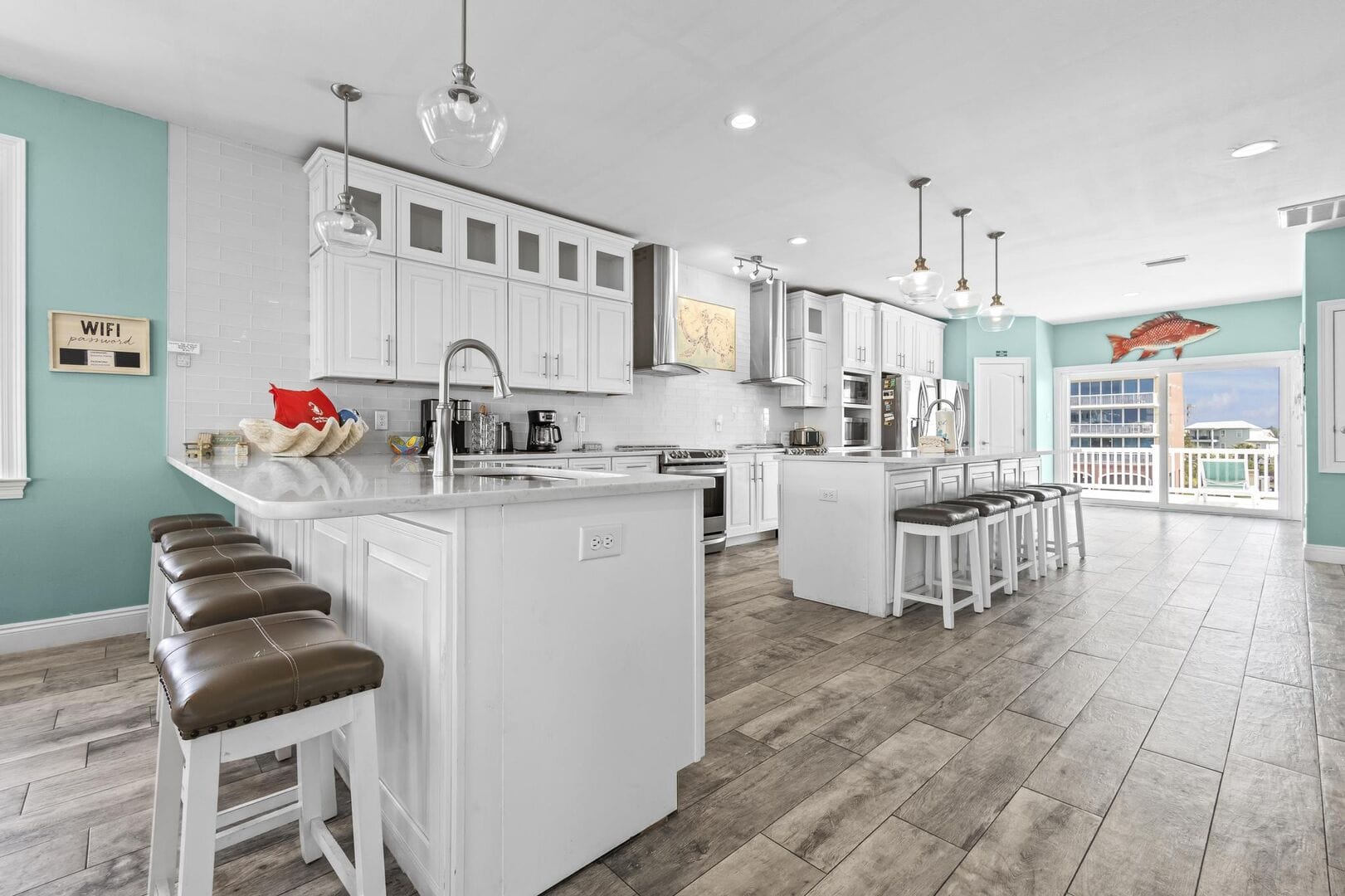 Spacious coastal kitchen with white cabinetry, two islands, and bar seating. Features include stainless steel appliances, pendant lighting, and large windows with balcony access and ocean views.