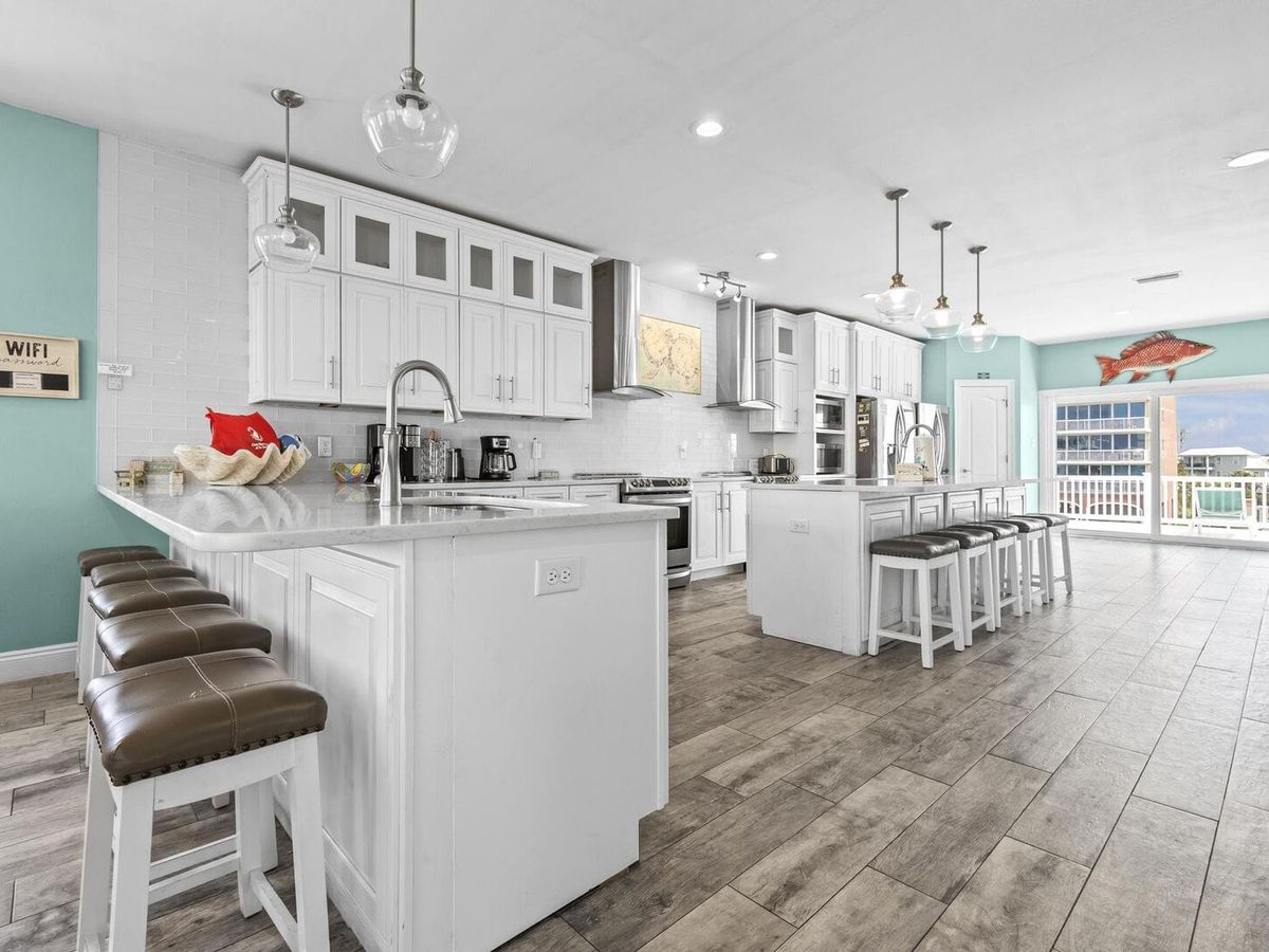Spacious coastal kitchen with white cabinetry, two islands, and bar seating. Features include stainless steel appliances, pendant lighting, and large windows with balcony access and ocean views.