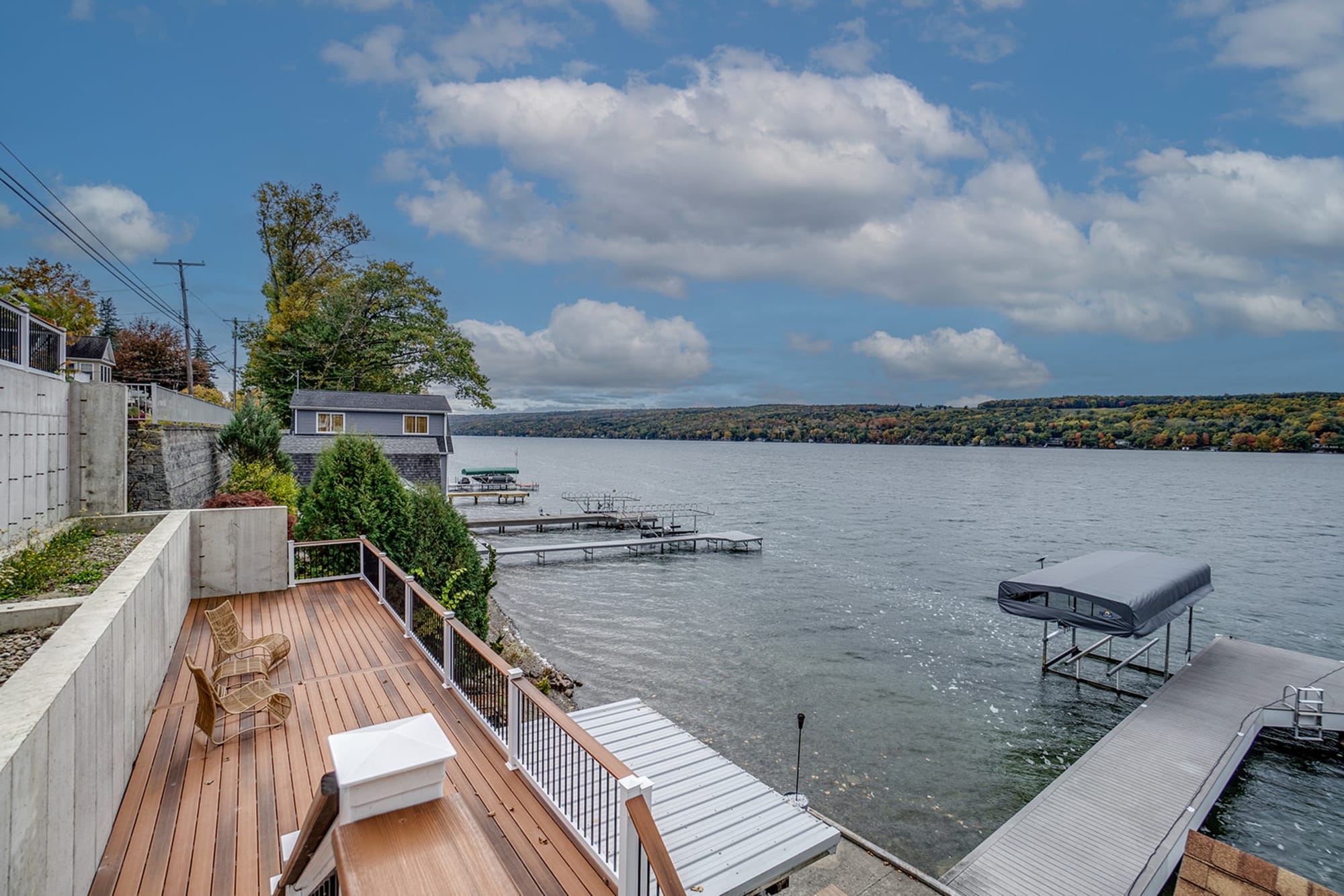 A scenic lakeside property featuring a wooden deck with seating, overlooking a calm lake with multiple docks extending into the water. A boathouse sits along the shoreline, while a tree-covered hillside with autumn foliage stretches across the horizon. 