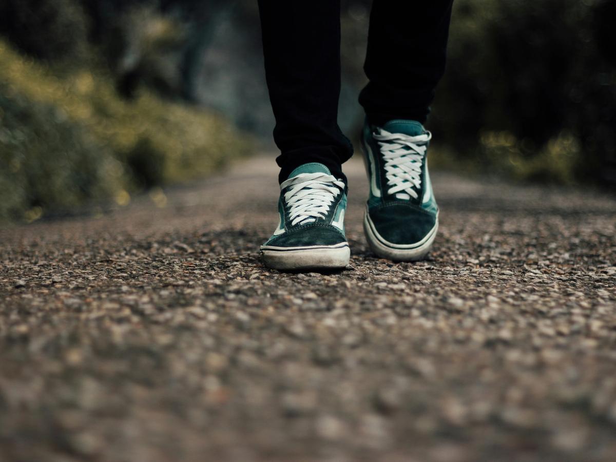 A close-up view of a person in black pants walking on a gravel path, wearing worn teal sneakers with white laces, surrounded by blurred greenery in the background.