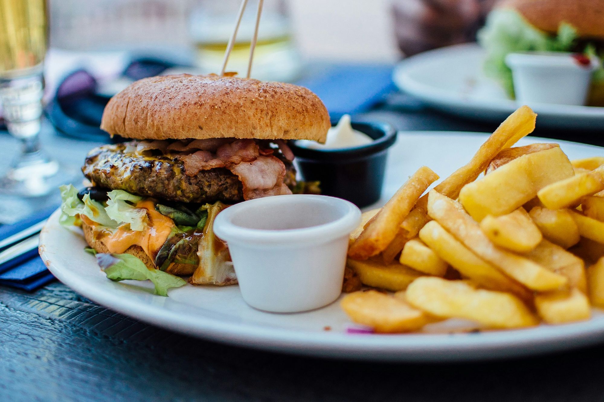 Close-up of a juicy bacon cheeseburger with lettuce and sauce, served with thick-cut golden fries and dipping sauces on a white plate.
