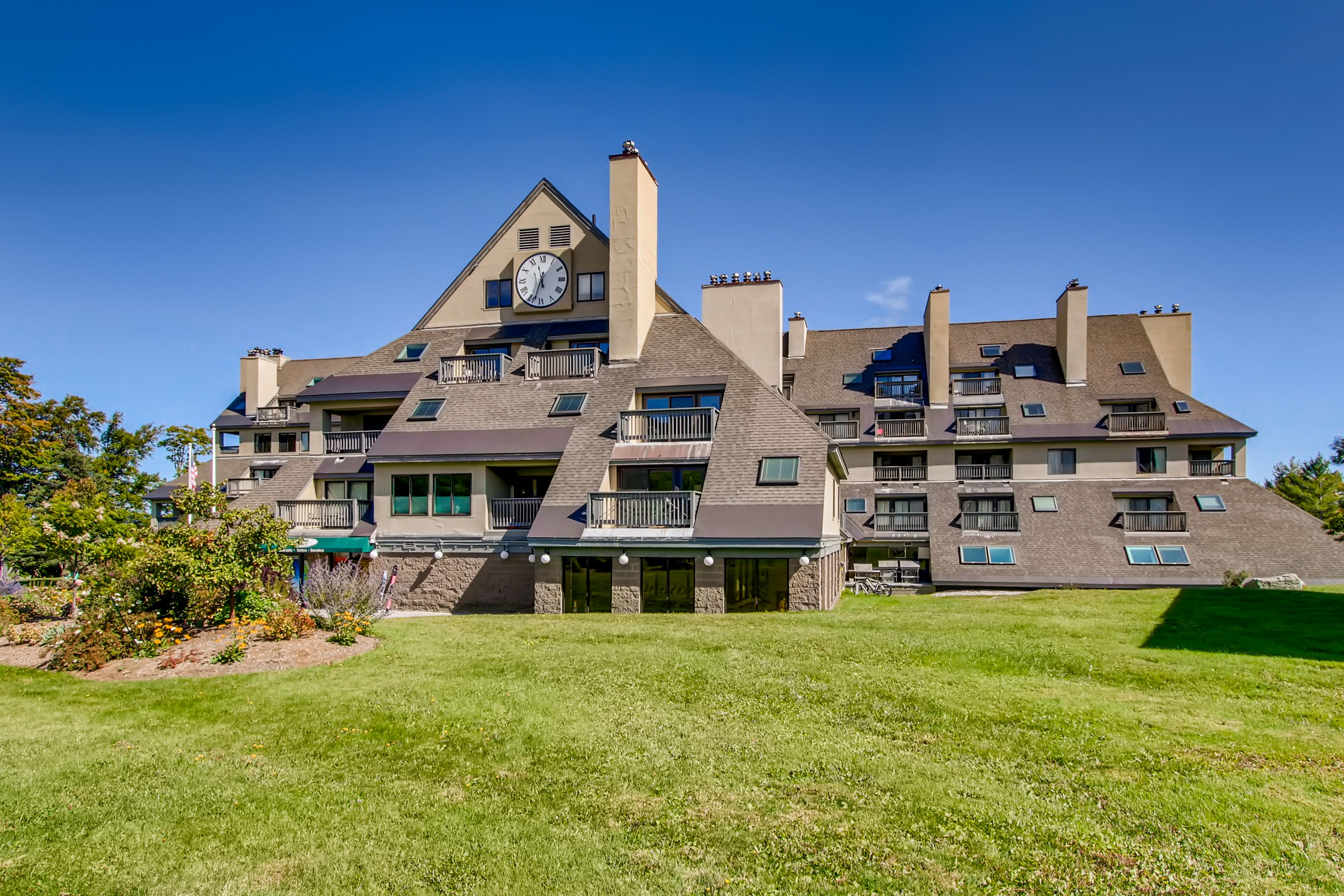 A large mountain resort building with sloped roofs, balconies, and a prominent clock tower, set against a clear blue sky with a manicured lawn and flower beds in the foreground.