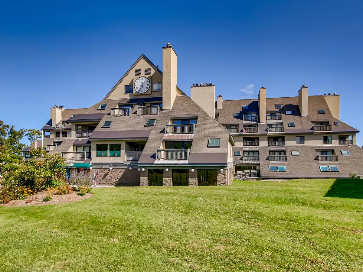A large mountain resort building with sloped roofs, balconies, and a prominent clock tower, set against a clear blue sky with a manicured lawn and flower beds in the foreground.