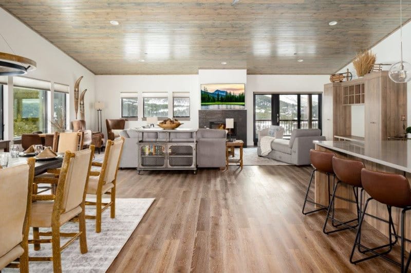 An open-concept mountain home with a modern rustic design features a dining area, cozy living room with fireplace, and kitchen with bar seating, all framed by large windows and scenic views.