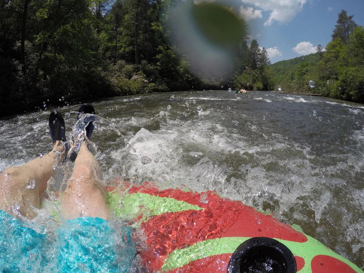 A person floats down a rushing river on a bright inflatable tube, feet splashing in the cool water. The forested banks and mountain backdrop create a fun and adventurous outdoor setting. Sunlight sparkles on the waves, making it a perfect summer activity.