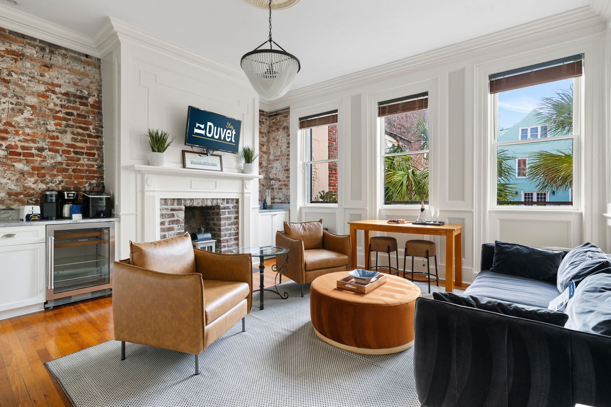 A chic living room features exposed brick walls, a white fireplace with a mounted TV, tan leather chairs, a velvet navy couch, and large windows with views of palm trees and colorful buildings.