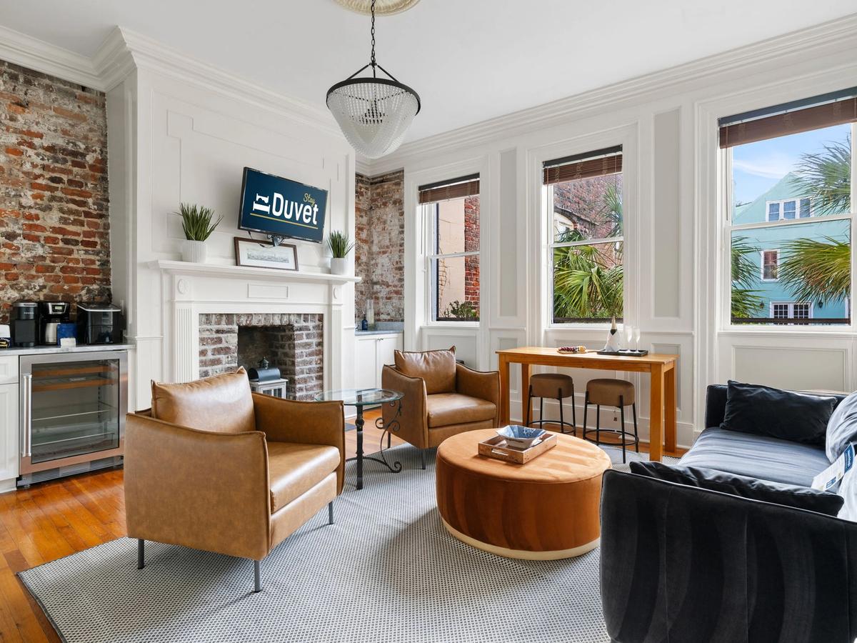 A chic living room features exposed brick walls, a white fireplace with a mounted TV, tan leather chairs, a velvet navy couch, and large windows with views of palm trees and colorful buildings.