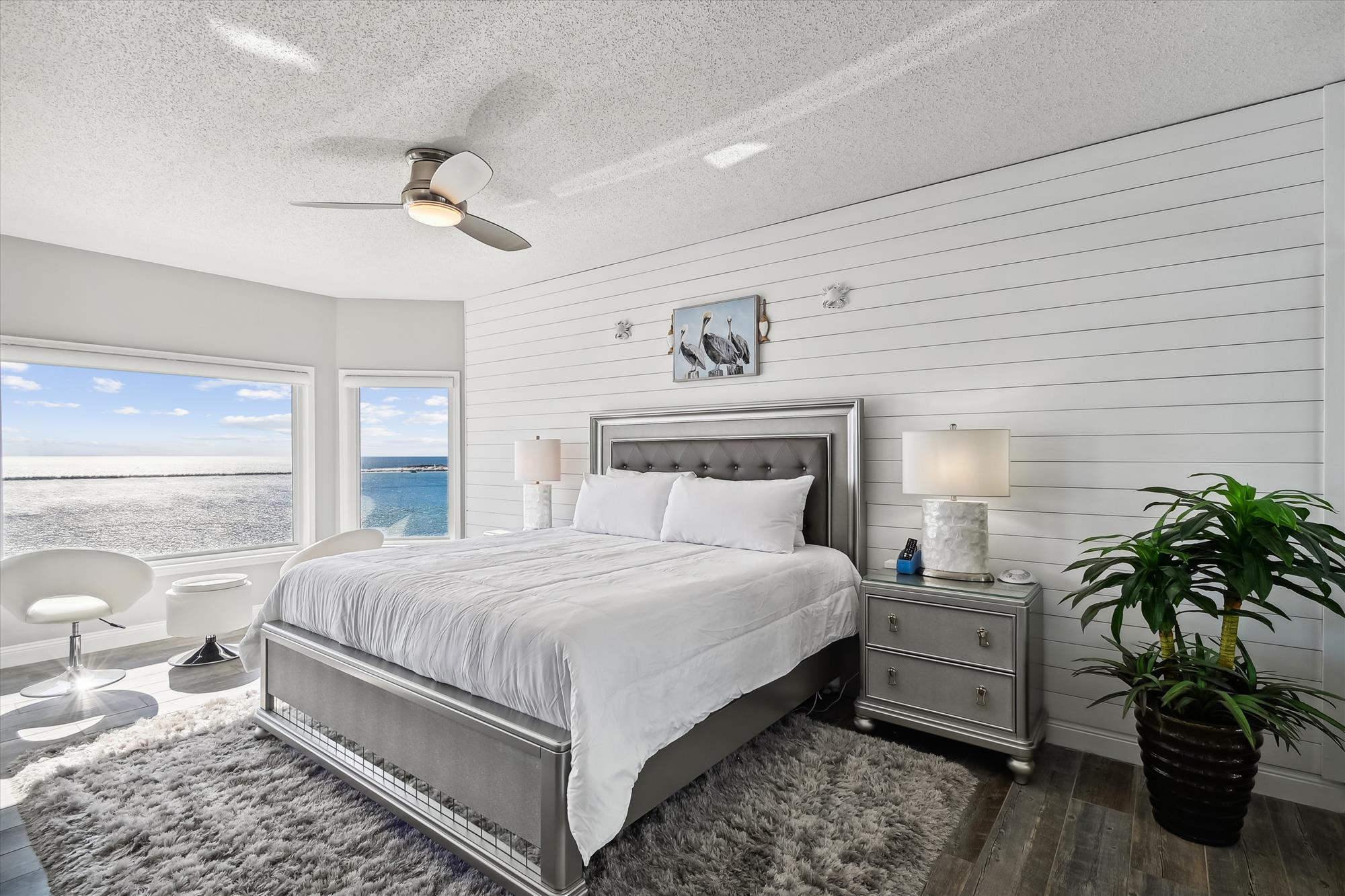 A bright, modern bedroom with a tufted gray bed, shiplap accent wall, ocean view windows, and sleek white furniture, all grounded by a plush gray rug and dark wood flooring.