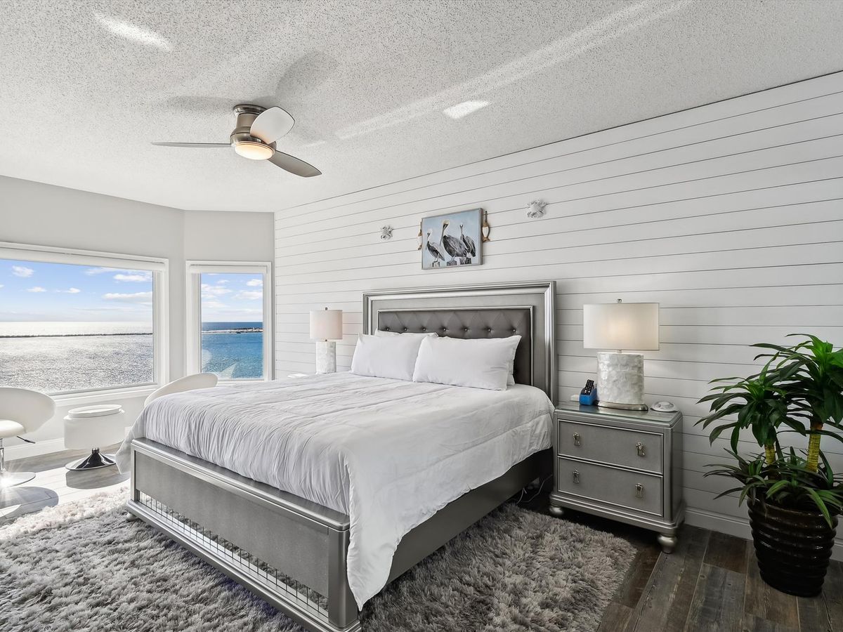 A bright, modern bedroom with a tufted gray bed, shiplap accent wall, ocean view windows, and sleek white furniture, all grounded by a plush gray rug and dark wood flooring.