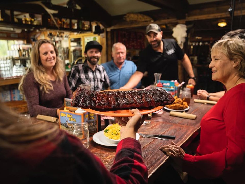Friends gather at Riverhouse BBQ in Big Sky with a rack of smoky ribs served family-style. A lively restaurant known for barbecue, beers, and mountain views.