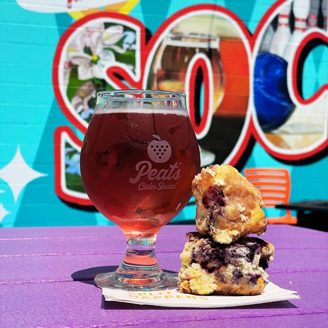 A glass of berry cider and blueberry pastries sit on a vibrant purple table in front of a colorful mural at Peat’s Cider Social. The playful backdrop and bright colors capture the fun, laid-back vibe of this local favorite.