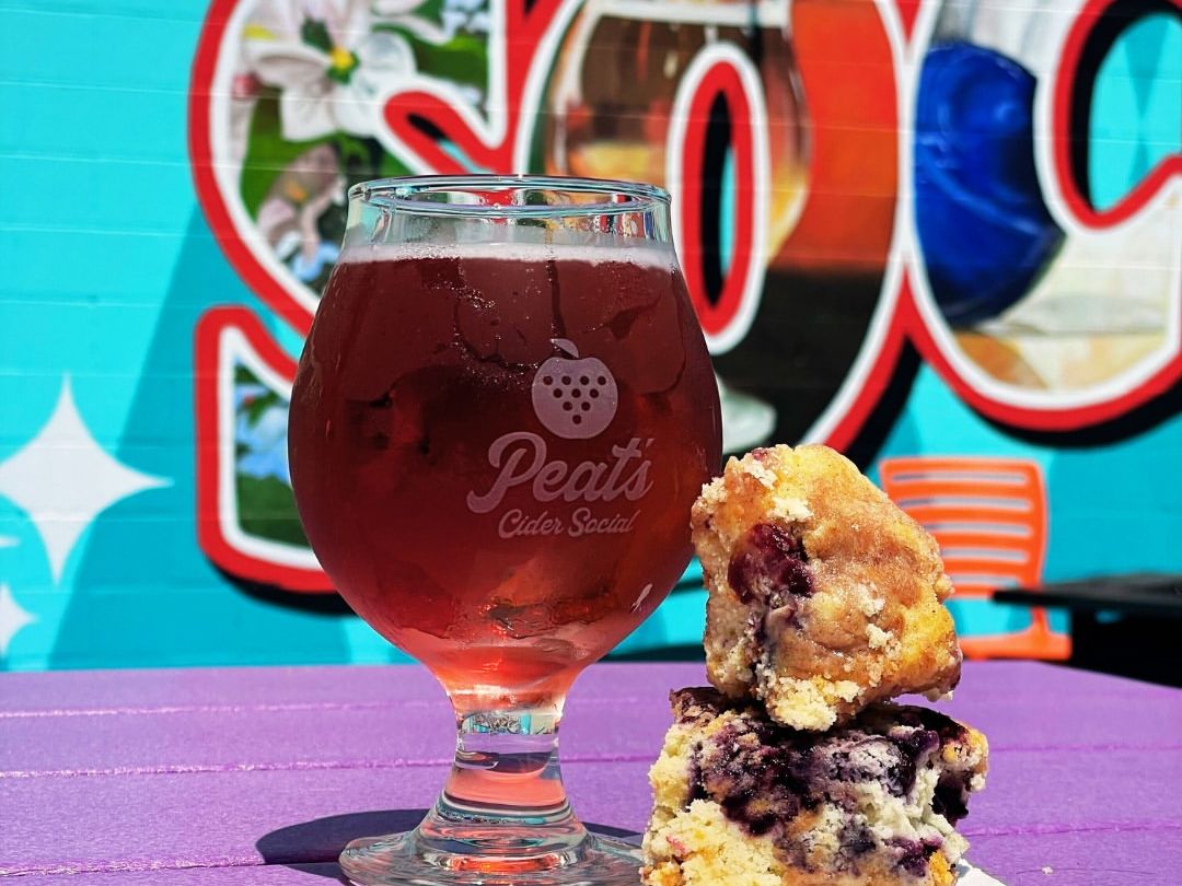 A glass of berry cider and blueberry pastries sit on a vibrant purple table in front of a colorful mural at Peat’s Cider Social. The playful backdrop and bright colors capture the fun, laid-back vibe of this local favorite.