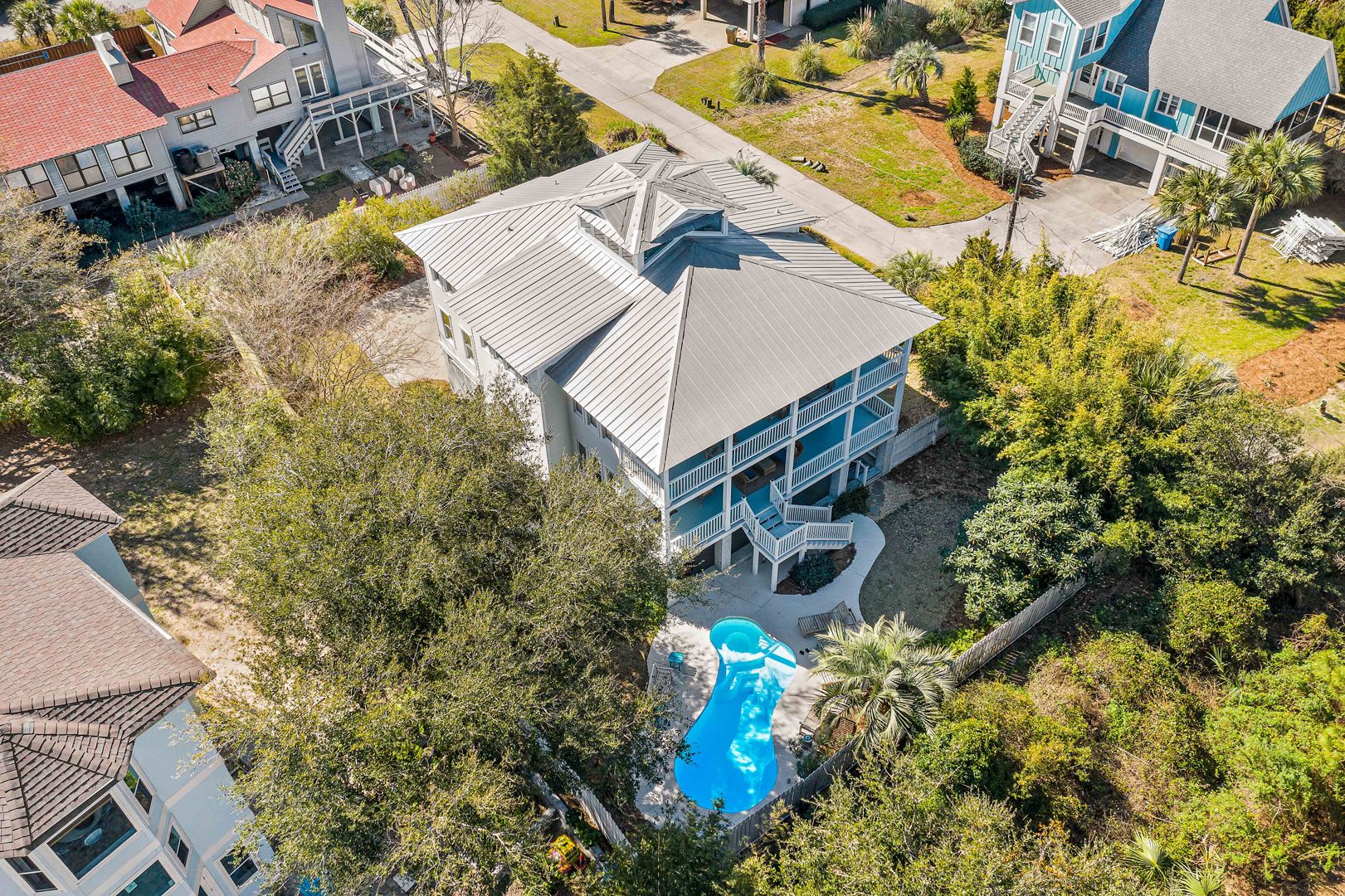 Aerial view of a coastal home with a gray metal roof and double wraparound porches, surrounded by trees and featuring a private backyard with a curvy pool and lush landscaping.