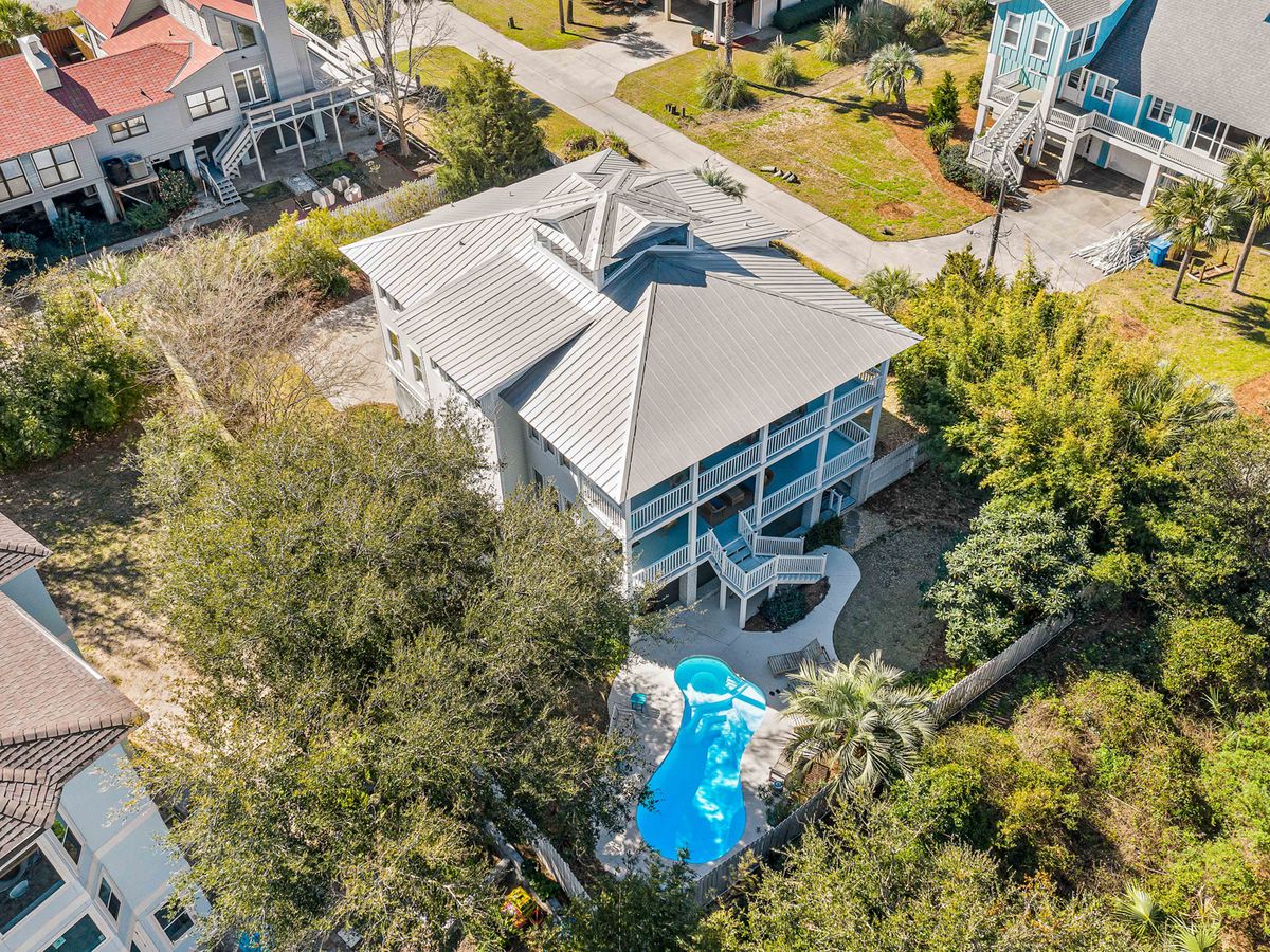 Aerial view of a coastal home with a gray metal roof and double wraparound porches, surrounded by trees and featuring a private backyard with a curvy pool and lush landscaping.