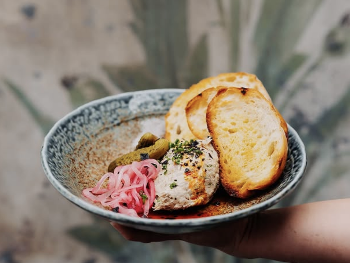 A rustic ceramic bowl holds a gourmet spread of toasted bread slices, a scoop of herbed tuna or crab salad, pickled red onions, and small pickles, presented against a blurred botanical background.