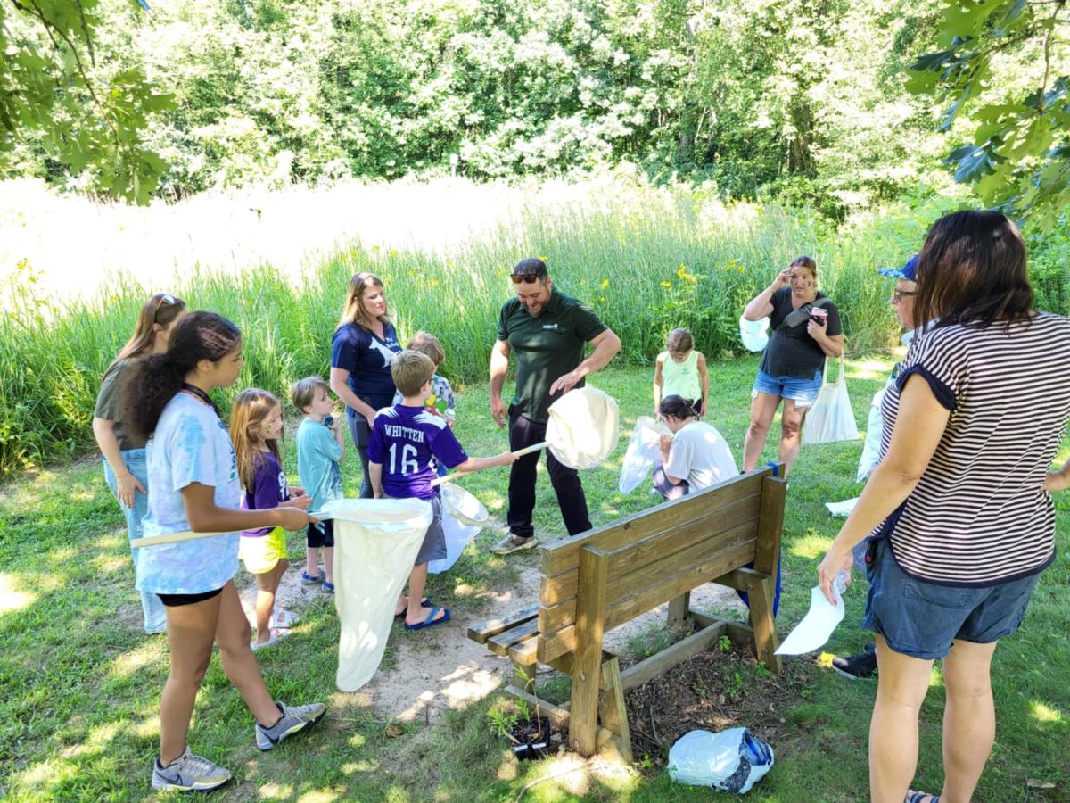 Families gather at Love Creek County Park and Nature Center to explore nature and learn together. Kids hold butterfly nets as they listen to a guide, surrounded by lush greenery and summer sunshine.
