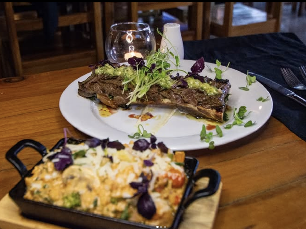 Gourmet dinner featuring a plate of roasted bone marrow topped with herbs and a side of cheesy vegetable casserole, served on a wooden table with a candle