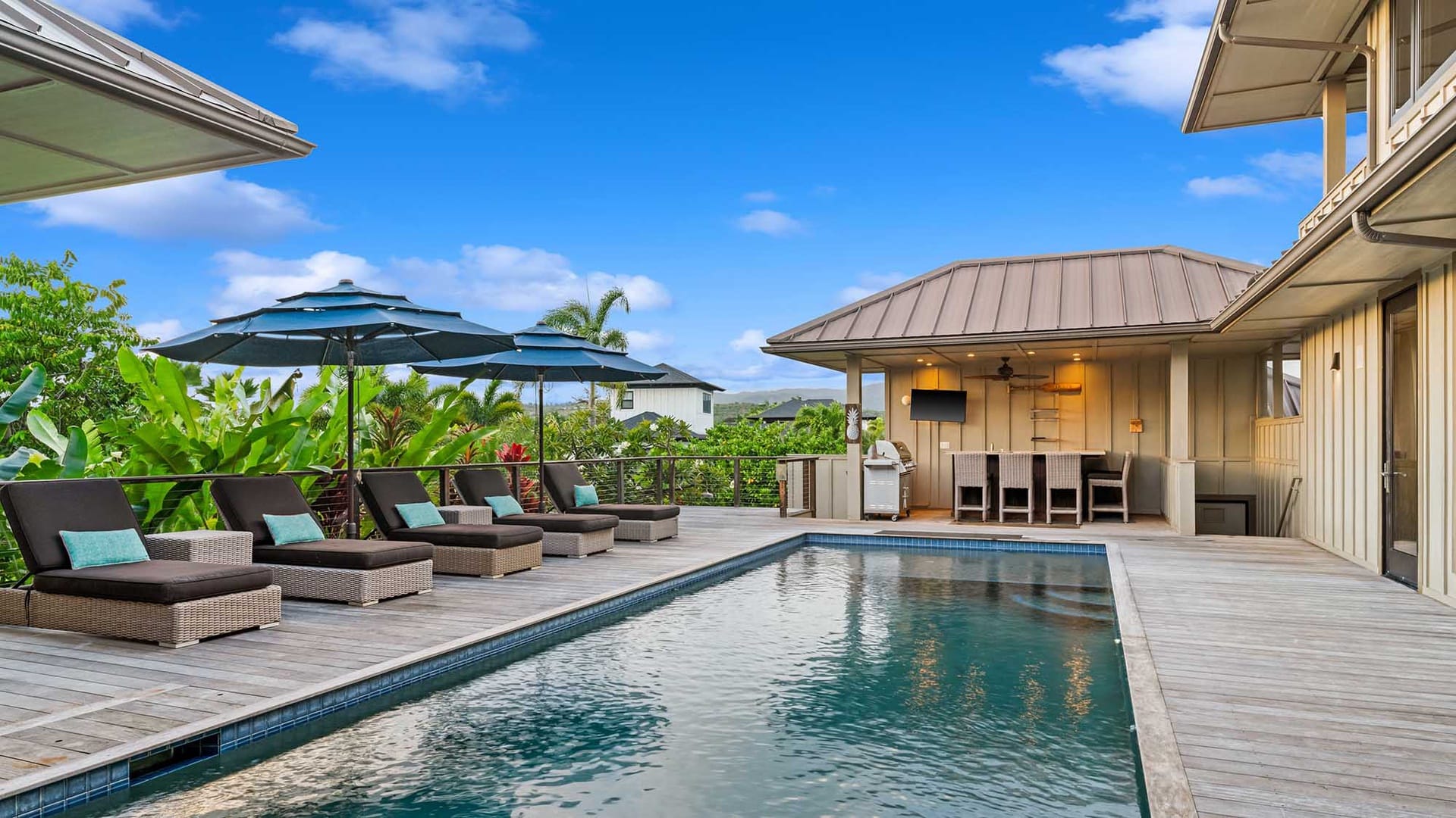 Modern tropical pool deck with lounge chairs, large umbrellas, and an outdoor bar area beside a sleek rectangular pool surrounded by lush greenery.