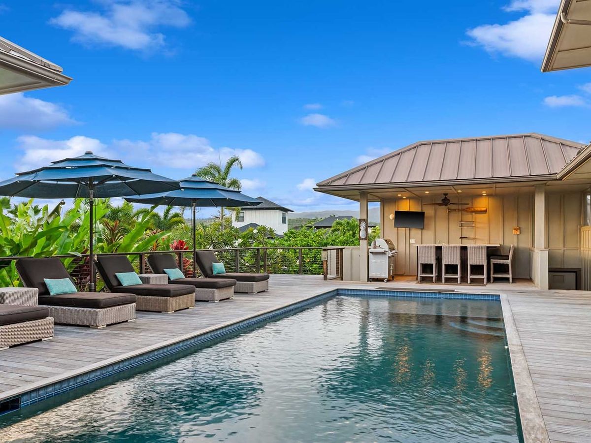Modern tropical pool deck with lounge chairs, large umbrellas, and an outdoor bar area beside a sleek rectangular pool surrounded by lush greenery.