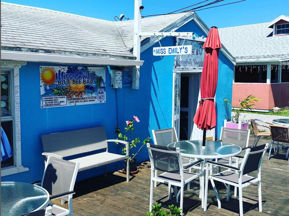 A bright blue building houses Miss Emily’s Blue Bee Bar, featuring outdoor patio seating with glass tables, a red umbrella, and tropical charm under a sunny Bahamian sky.