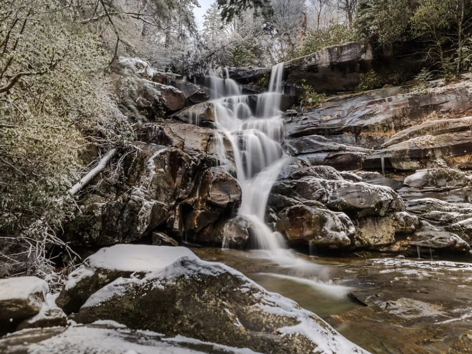 A cascading waterfall flows over layered rocks dusted with snow and ice, surrounded by frosted foliage in a serene winter setting in the Great Smoky Mountains.