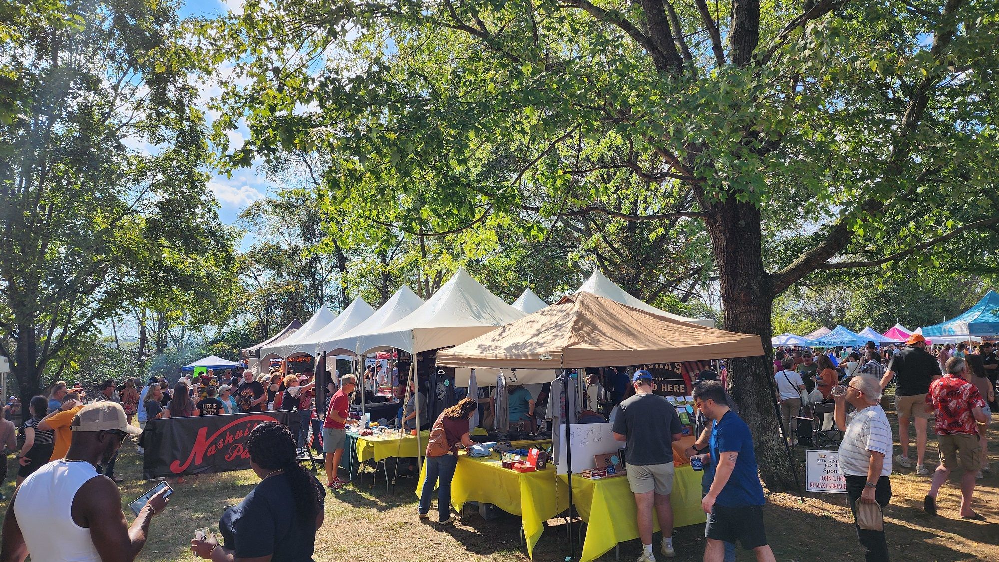 Crowds gather under rows of festival tents, enjoying drinks, food, and sunny weather at the Tennessee Beer, Wine, and Shine Festival. People chat, taste samples, and explore local vendors under the big shade trees. The whole scene has a lively, friendly vibe perfect for a fall day out.