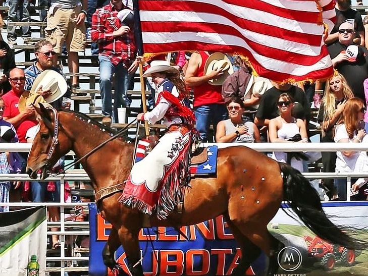 A rodeo performer waves a large American flag on horseback as the crowd cheers. The Red Lodge Rodeo brings Western tradition to life.