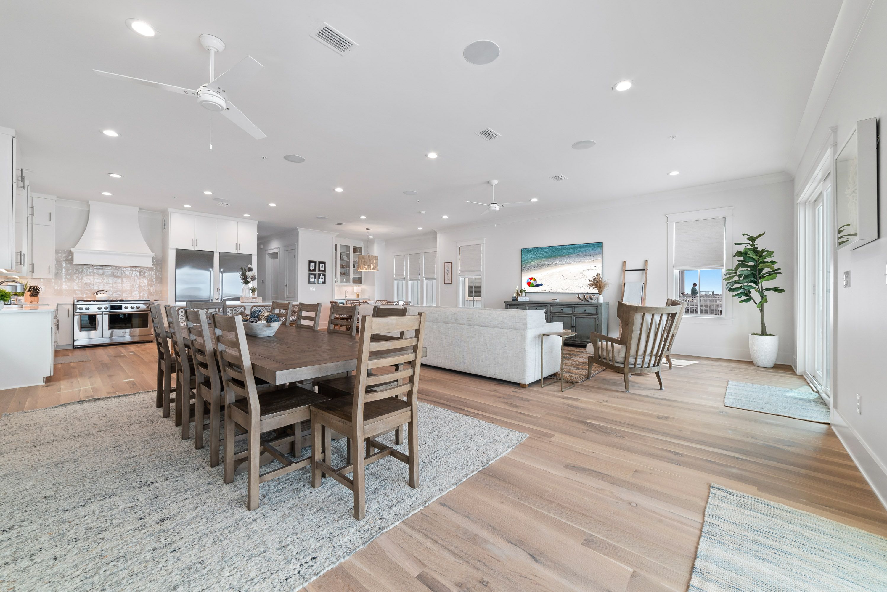 A spacious open-concept coastal home interior with a large wood dining table, modern white kitchen, cozy living area, light wood flooring, and bright natural light from multiple windows and glass doors.