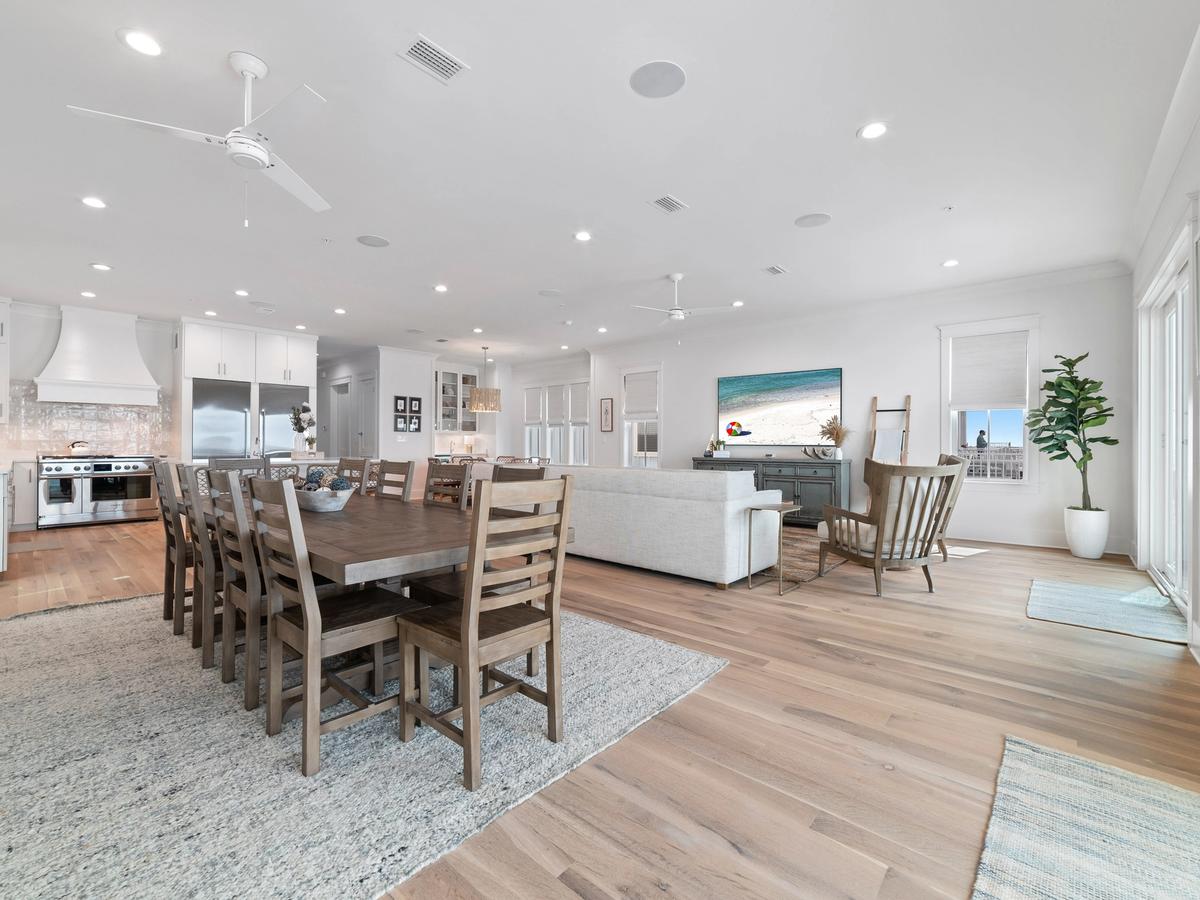 A spacious open-concept coastal home interior with a large wood dining table, modern white kitchen, cozy living area, light wood flooring, and bright natural light from multiple windows and glass doors.