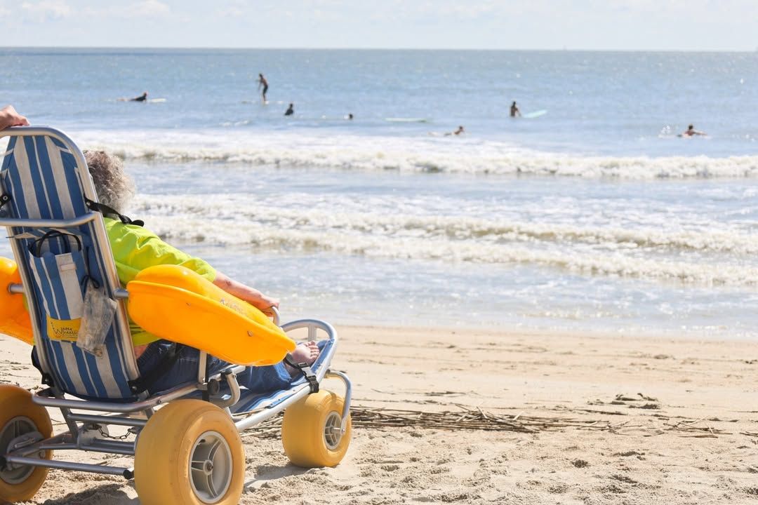 An adaptive beach wheelchair rests near the shoreline as visitors surf and swim in the background. The scene reflects Tybee’s welcoming atmosphere and commitment to making the ocean accessible to everyone.