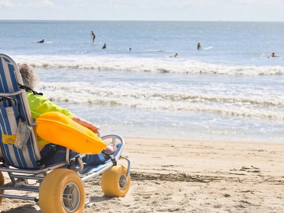 An adaptive beach wheelchair rests near the shoreline as visitors surf and swim in the background. The scene reflects Tybee’s welcoming atmosphere and commitment to making the ocean accessible to everyone.