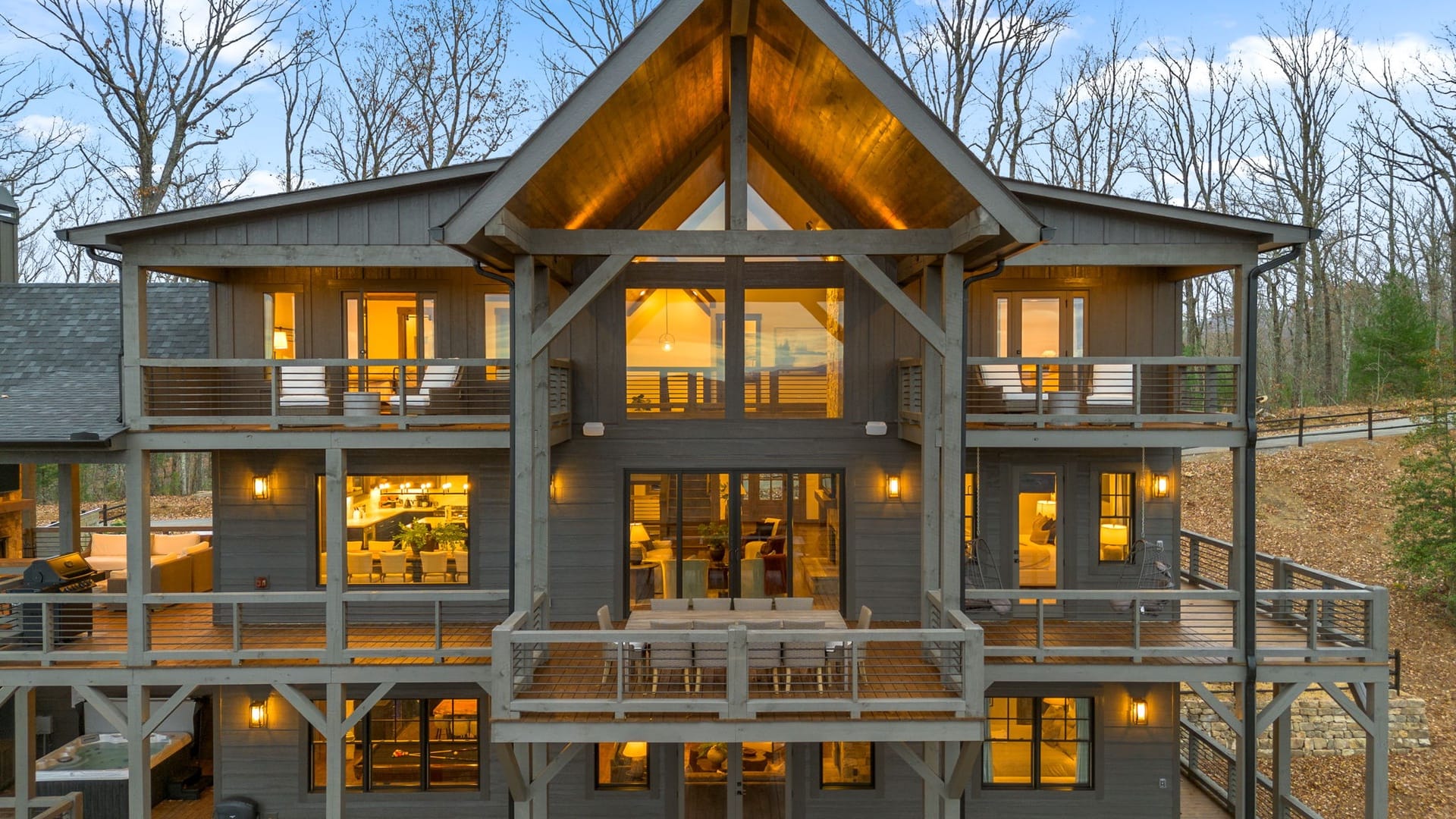 A three-story modern mountain cabin with expansive wraparound decks, exposed timber framing, and warm interior lighting glowing through large windows, set among leafless trees at dusk.