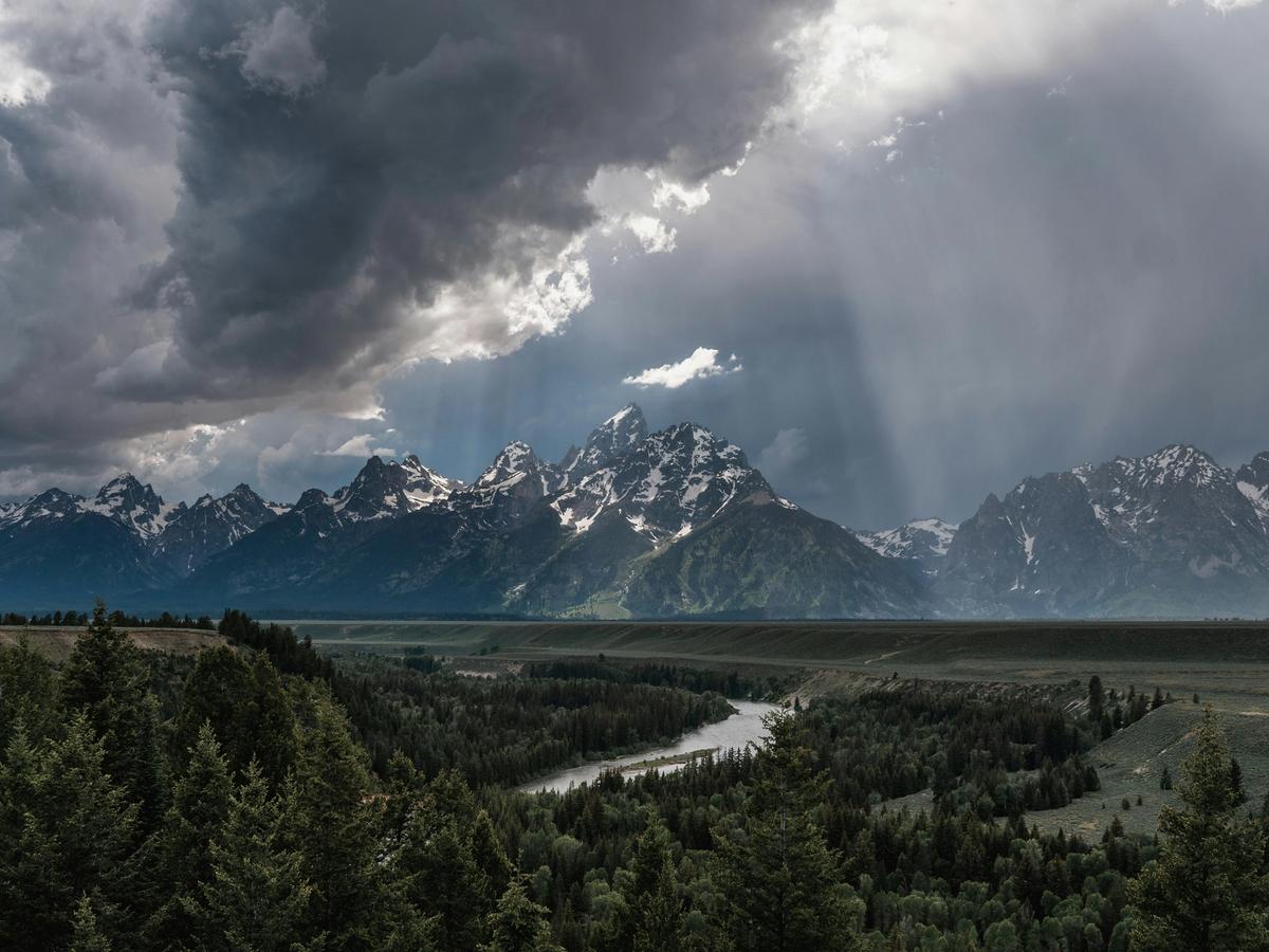 Dramatic storm clouds and rays of light pierce the sky above the rugged Teton Range, casting shadows over the Snake River as it winds through a lush, forested valley.