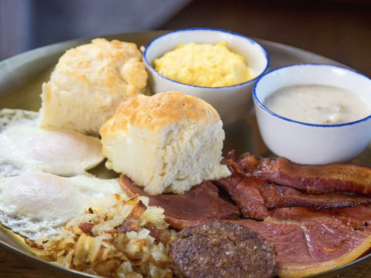 A hearty Southern breakfast plate featuring sunny-side-up eggs, hash browns, crispy bacon, country ham, sausage, two fluffy biscuits, and small bowls of scrambled eggs and sausage gravy.