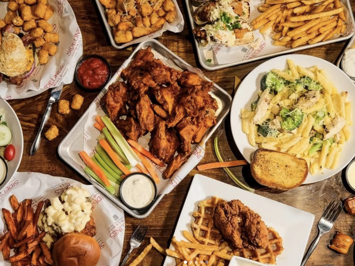 A hearty spread of American comfort food includes trays of buffalo wings with celery and ranch, chicken and waffles, mac and cheese-topped burgers, tacos with fries, pasta with broccoli and garlic bread, and baskets of tater tots, all served on branded "Sidelines" paper.