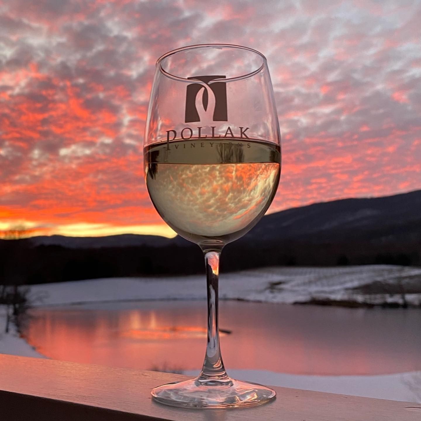 A glass of white wine from Pollak Vineyards glows against a vibrant sunset over rolling hills and a reflective pond. The warm colors and tranquil view capture the romantic beauty of Virginia’s wine country.