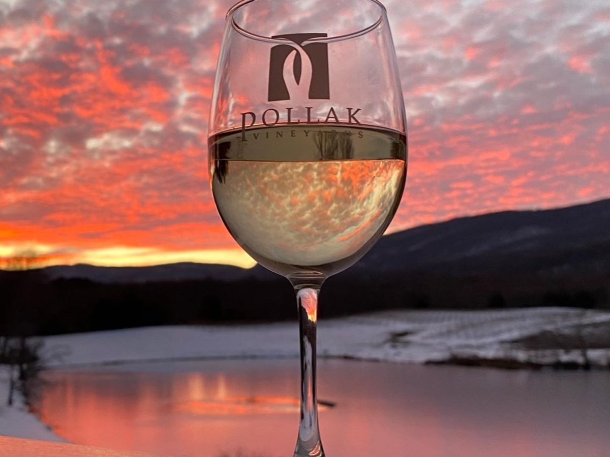 A glass of white wine from Pollak Vineyards glows against a vibrant sunset over rolling hills and a reflective pond. The warm colors and tranquil view capture the romantic beauty of Virginia’s wine country.
