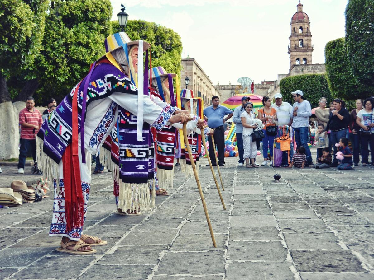 Traditional Mexican dancers perform in colorful woven costumes and masks in a historic town square. A crowd of families watches as the performers move across the stone street near a colonial church. This vibrant cultural festival shows the rich heritage and local traditions of Mexico.