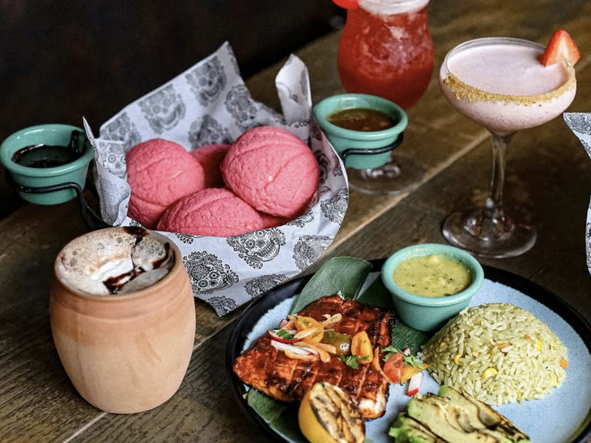 A colorful Mexican-inspired meal with grilled fish, rice, avocado, pink conchas, dipping sauces, and vibrant cocktails on a rustic wooden table.