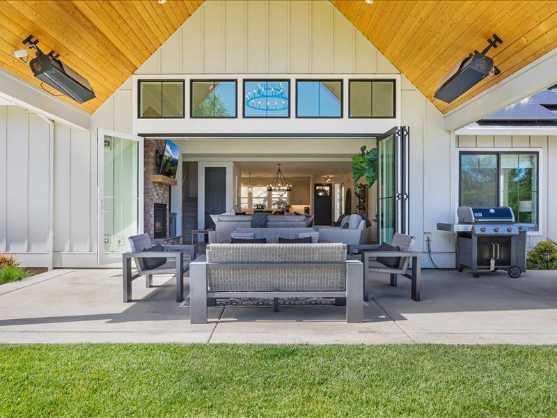 A modern covered patio with outdoor seating opens seamlessly into a spacious living room, featuring a mounted TV, ceiling heaters, a barbecue grill, and a high wood-paneled ceiling.