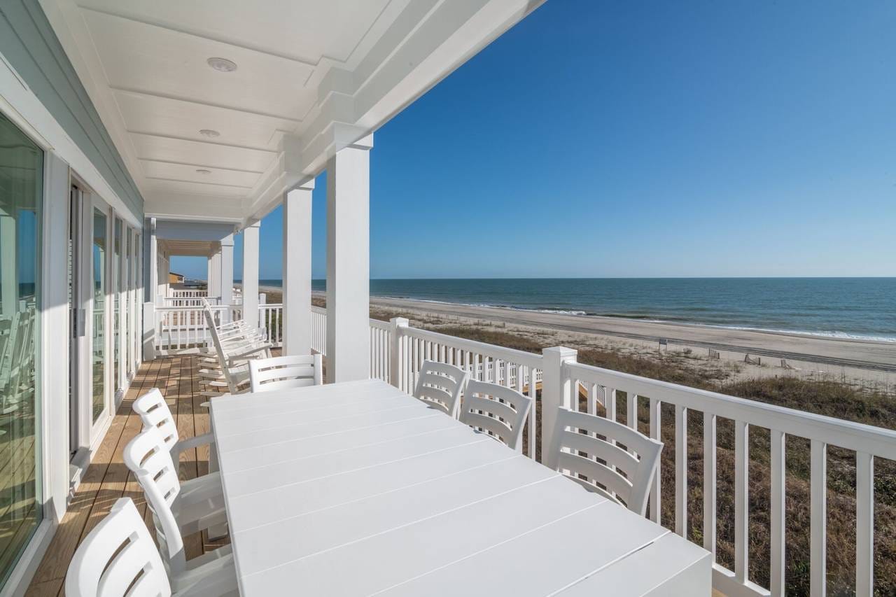 A spacious beachfront balcony with white railings and columns, overlooking a sandy shore and the ocean. A long white dining table with matching chairs sits in the foreground, while several white rocking chairs line the wooden deck near large sliding glass doors. The clear blue sky and calm waves create a serene coastal view.