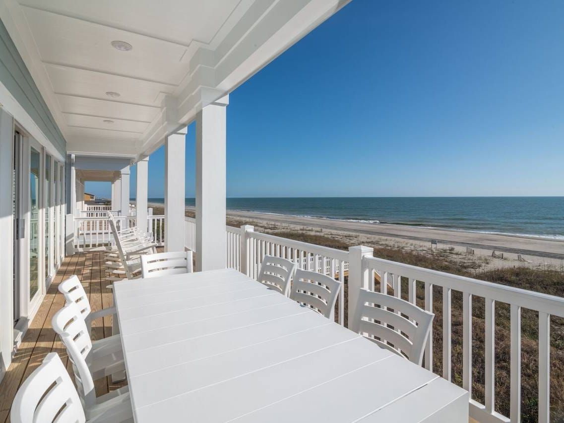 A spacious beachfront balcony with white railings and columns, overlooking a sandy shore and the ocean. A long white dining table with matching chairs sits in the foreground, while several white rocking chairs line the wooden deck near large sliding glass doors. The clear blue sky and calm waves create a serene coastal view.