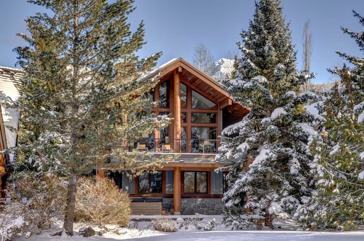 A ski chalet in Whistler, BC, nestled among tall snow-covered evergreen trees with a snow-capped mountain peak visible in the background on a bright winter day. The cabin features a striking A-frame roofline, exposed log timber beams, floor-to-ceiling windows, and a mix of stone and dark-panel siding that fits right into the mountain landscape.