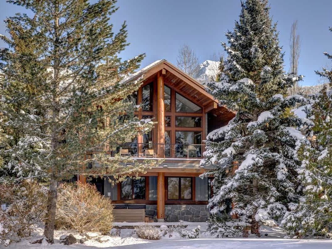 A ski chalet in Whistler, BC, nestled among tall snow-covered evergreen trees with a snow-capped mountain peak visible in the background on a bright winter day. The cabin features a striking A-frame roofline, exposed log timber beams, floor-to-ceiling windows, and a mix of stone and dark-panel siding that fits right into the mountain landscape.