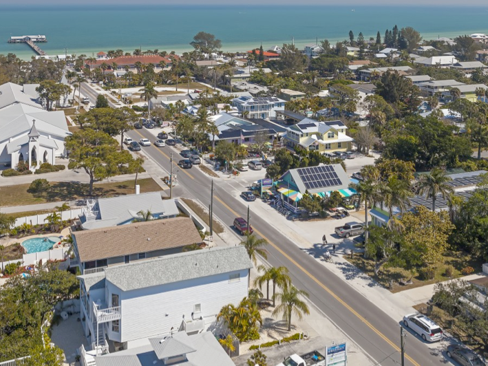 Aerial view of the Anna Maria Historic Green Village on Florida’s Gulf Coast, featuring eco-friendly buildings with solar panels, colorful umbrellas, and a charming small-town vibe just steps from the beach. The turquoise water and iconic pier can be seen in the background, showing off the laid-back charm of Anna Maria Island.