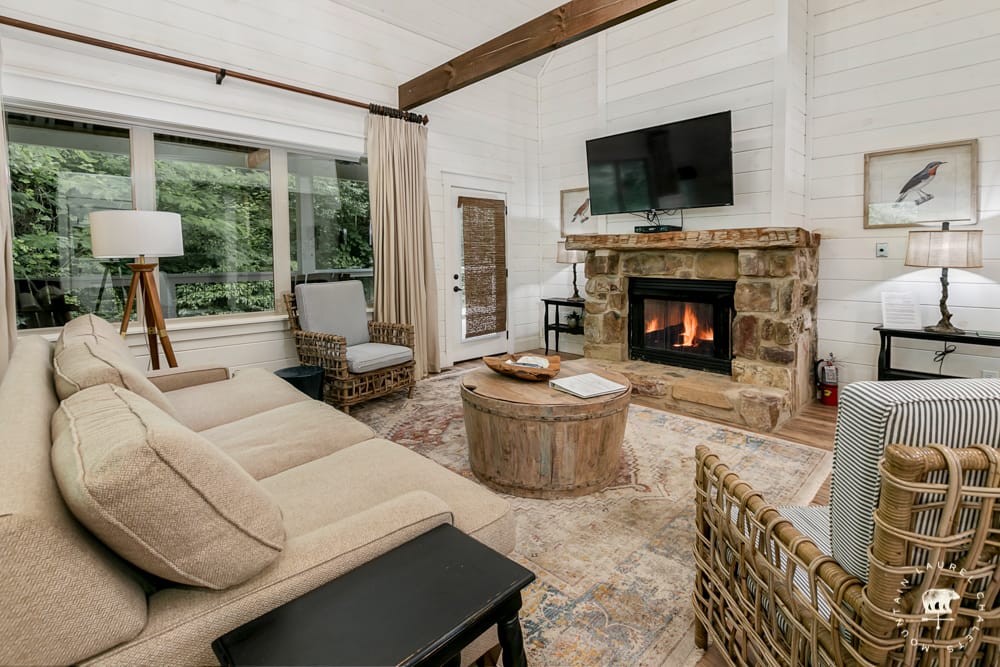 A cozy living room with a stone fireplace and mounted TV features a beige sectional sofa, wicker chairs, and a rustic wooden coffee table, accented by large windows that bring in views of the greenery outside.