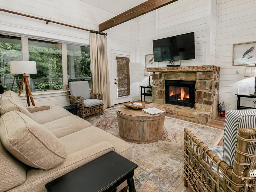 A cozy living room with a stone fireplace and mounted TV features a beige sectional sofa, wicker chairs, and a rustic wooden coffee table, accented by large windows that bring in views of the greenery outside.