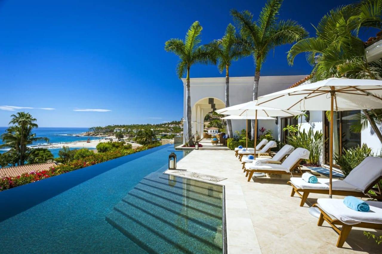 This infinity pool looks out over a bright blue coastline, with palm trees and white umbrellas creating a calm, sunny vibe. The scene feels like a perfect beach escape, where you can relax on a lounge chair and enjoy the ocean breeze. It captures the beauty of a luxury villa set high above the shore.