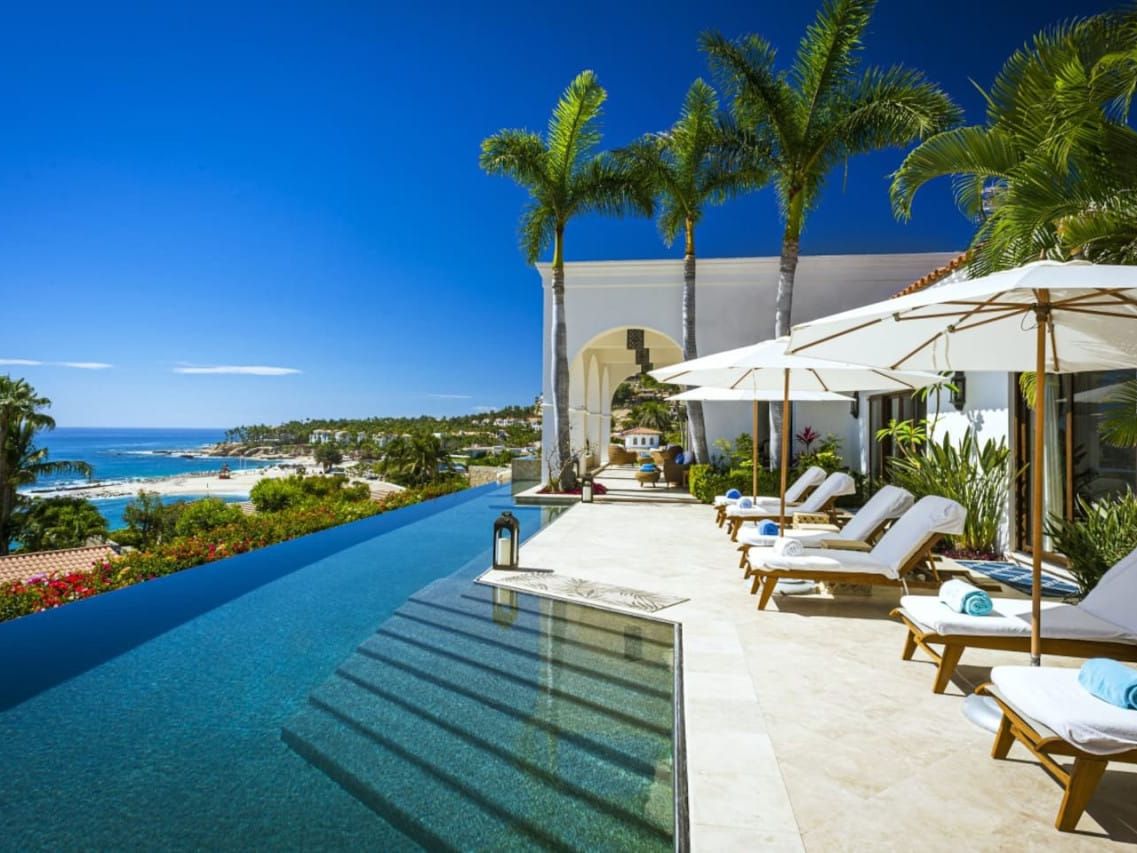 This infinity pool looks out over a bright blue coastline, with palm trees and white umbrellas creating a calm, sunny vibe. The scene feels like a perfect beach escape, where you can relax on a lounge chair and enjoy the ocean breeze. It captures the beauty of a luxury villa set high above the shore.